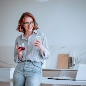 Portrait of a happy attractive woman using mobile phone while holding cup of tea and leaning on a table with laptop computer at home