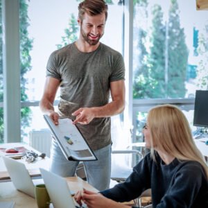 Image of smiling young colleagues sitting in office coworking using laptop computers and work with diagrama. Looking aside.